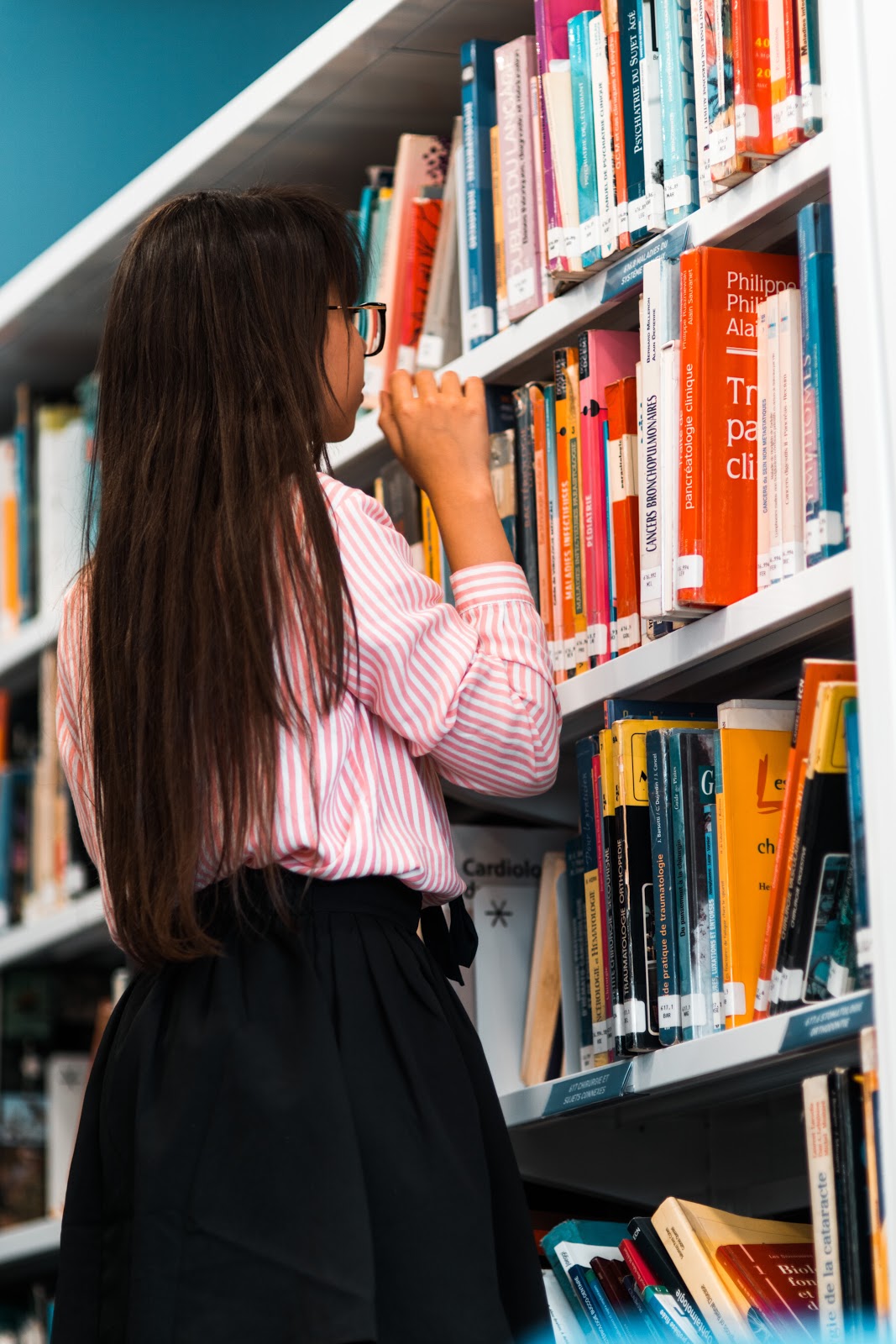 A young girl faces a shelf full of books at the library and appears to be choosing one.