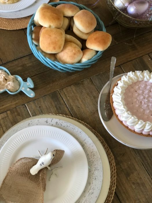 A wooden table with a wicker bowl with buns, a pie and a table setting with Easter decorations.