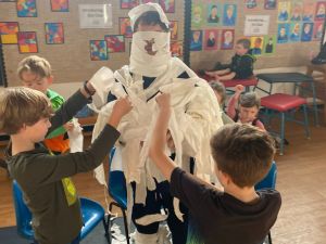 Children playfully dressing the sports coach as a Halloween Mummy using rolls of toilet paper.