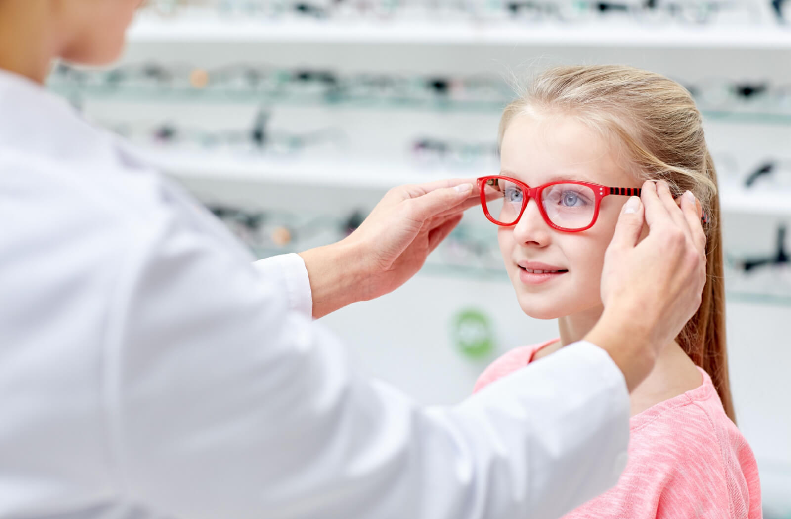 A child smiling and trying on glasses in an optical clinic while being assisted by an optometrist