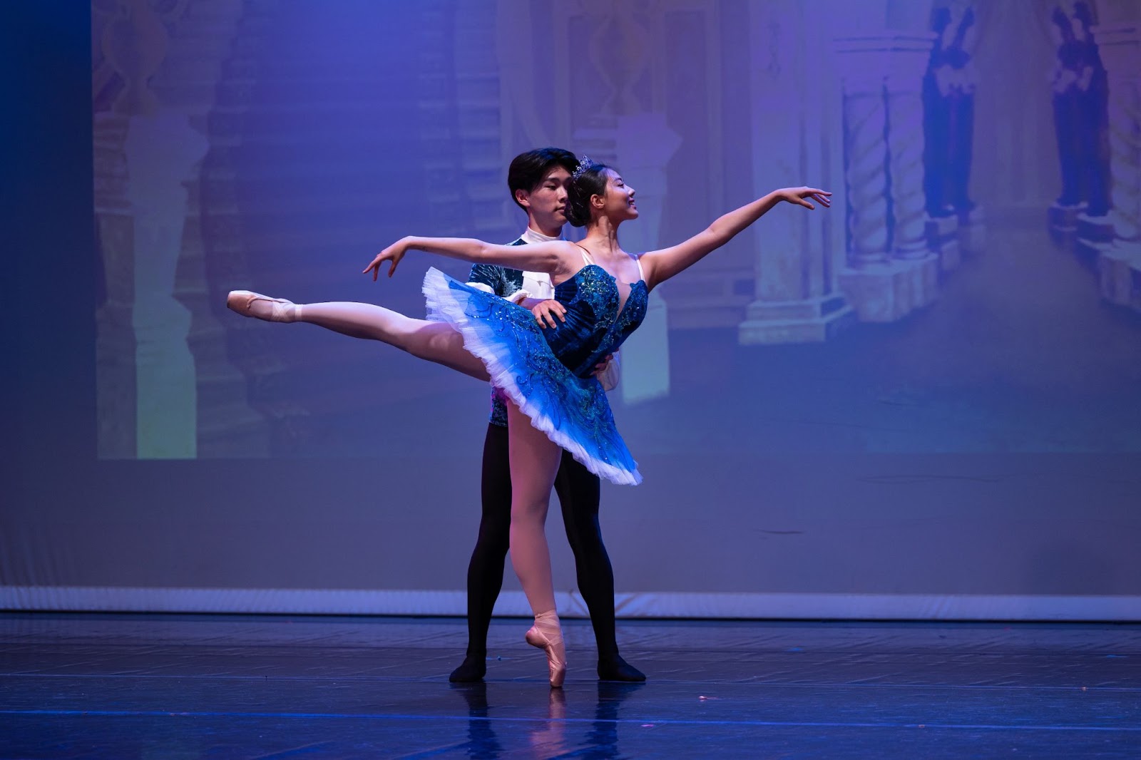 On stage, a male ballet dancer with black tights stands behind a female ballet dancer in a blue tutu. The female dancer stands on the toes of a single foot, with her hands gracefully outstretched forward and backward and her other leg straight horizontal.