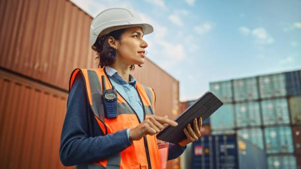 Smiling Portrait of a Beautiful Latin Female Industrial Engineer in White Hard Hat, High-Visibility Vest Working on Tablet Computer. Inspector or Safety Supervisor in Container Terminal. Smiling Portrait of a Beautiful Latin Female Industrial Engineer in White Hard Hat, High-Visibility Vest Working on Tablet Computer. Inspector or Safety Supervisor in Container Terminal. shipping container storage stock pictures, royalty-free photos & images