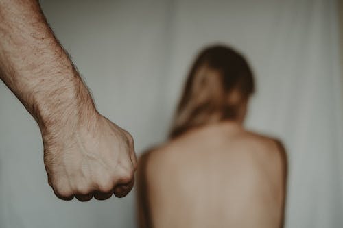 Free Aggressive abusive anonymous man clenching fist while insulting unrecognizable vulnerable female sitting on blurred background in light room during violence Stock Photo