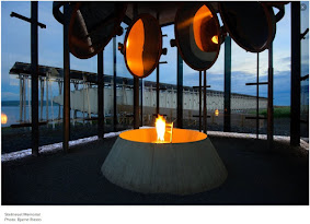 Steilneset memorial in the Norwegian arctic. Foreground, an art installation of a flaming chair, with large mirrored discs overhead reflecting the firelight. Background, a long hallway-like building with windows set in the side: 91 windows, one for each victim of the witchcraft trials here.