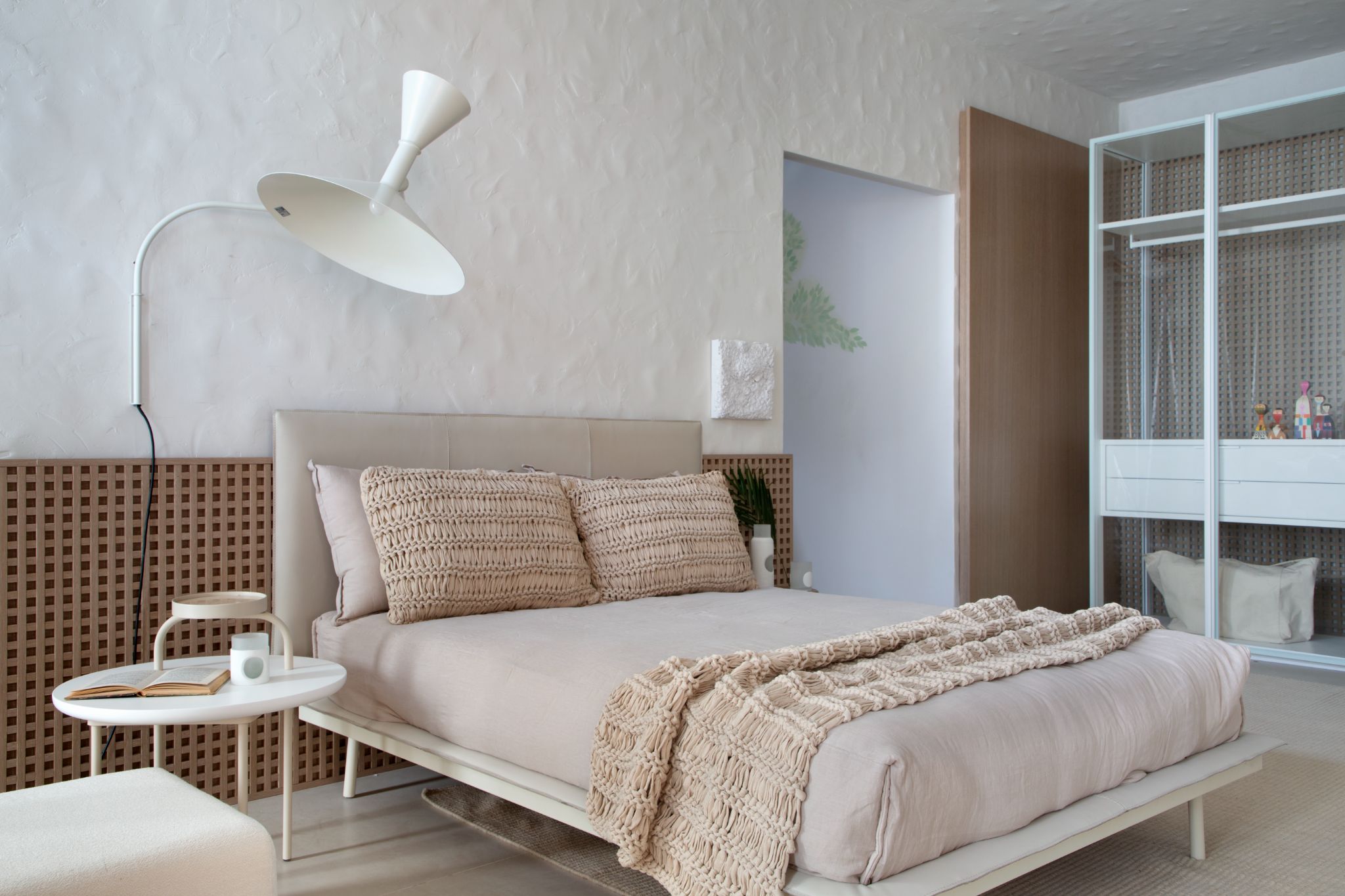 Quarto com cama de casal, elemento vazado em meia parede de fundo da cabeceira, piso branco, abajur branco e guarda roupa branco.