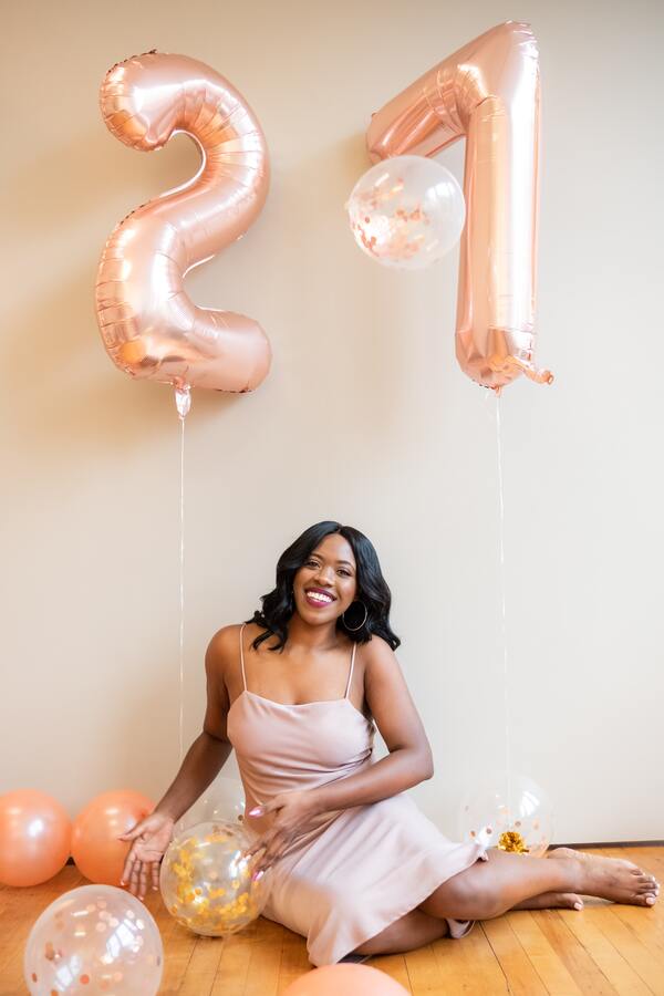 foto de uma mulher negra sentada no chão com bexigas e dois balões de letras formando 21