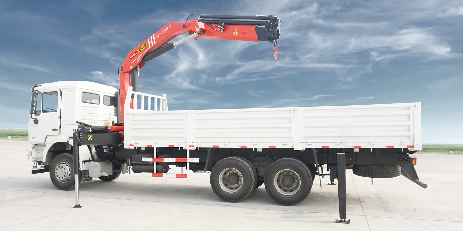 A truck mounted crane with white car paint and a red crane mounts its crane, and extend four outriggers several inches above the ground. The truck is parking on the ground under the blue sky, and showing its sideview.