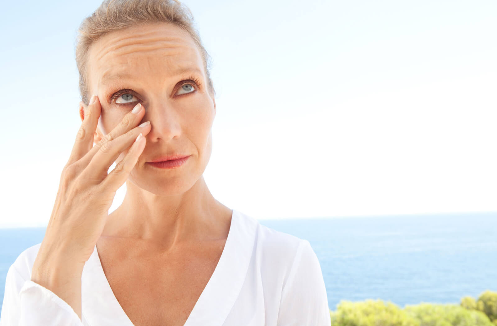 A woman in a white coloured blouse with her eyes getting dry while she is outside near the beachfront.