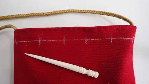 The bag is laid flat on a table and a white bone awl is laid out on top of it. One line spanning the width of the bag is marked in thin chalk 1/2" from the top and there are 6 lines marking spots for drawstring holes at regular intervals, drawn perpendicular to the first line.