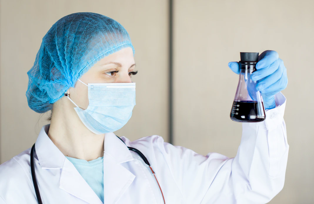 nurse in a blue knit cap and white lab coat holding a beaker with a purple liquid inside of it