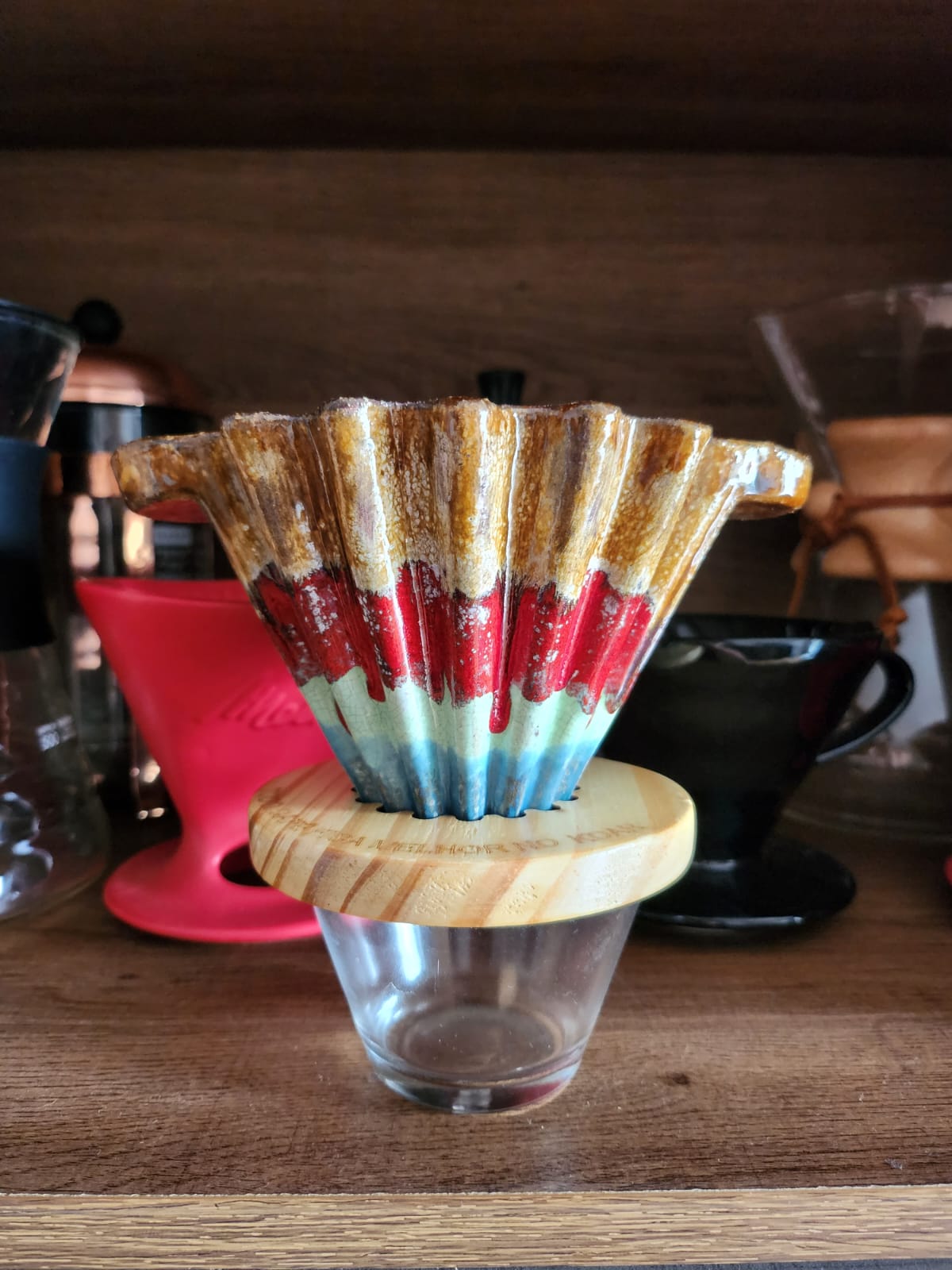 Foto de filtro Koar, feito em cerâmica. O filtro possui quatro cores diferentes em padrão reto, formando camadas de cima para baixo. Cores marrom, vermelho, azul claro e azul médio. A cor entre as camadas tem transição orgânica, com ângulos arredondados, como se as tintas entrassem em contato brevemente, porém sem se misturar.
Imagem com filtro Koar em destaque, com fundo de madeira. Outros métodos diversos de café ao fundo, desfocados.