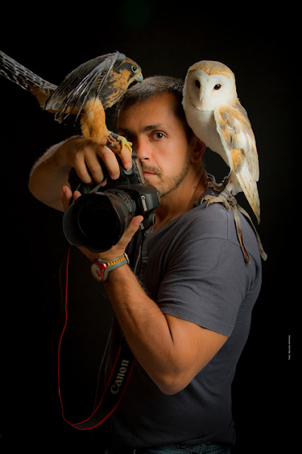 Retrato de Johnny Duarte. Ele  olha para frente, munido de sua câmera Canon e carrega duas aves nos ombros. Da esquerda para a direita, um falcão-de-coleira e uma coruja-das-torres.