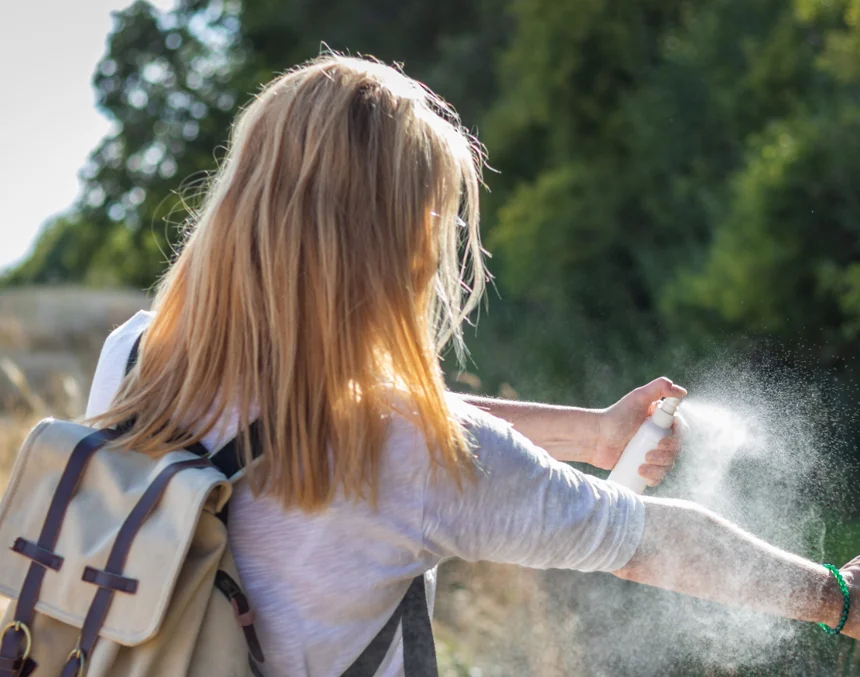 A young girl with blonde hair is spraying bug spray on her arm.