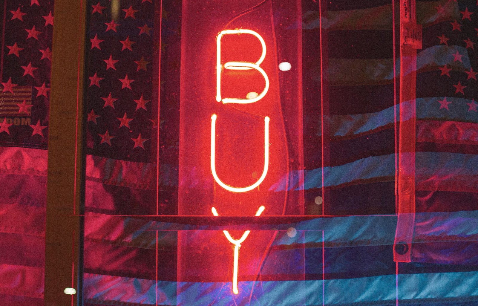 A red neon sign with the word BUY displayed vertically against an American flag.