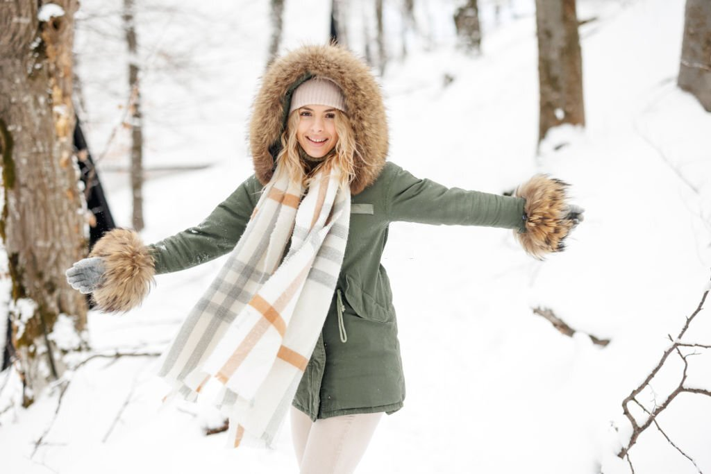 Beautiful young woman ina classy winter outfit hooded jacket standing with arms raised in snowy forest on cold winter day.