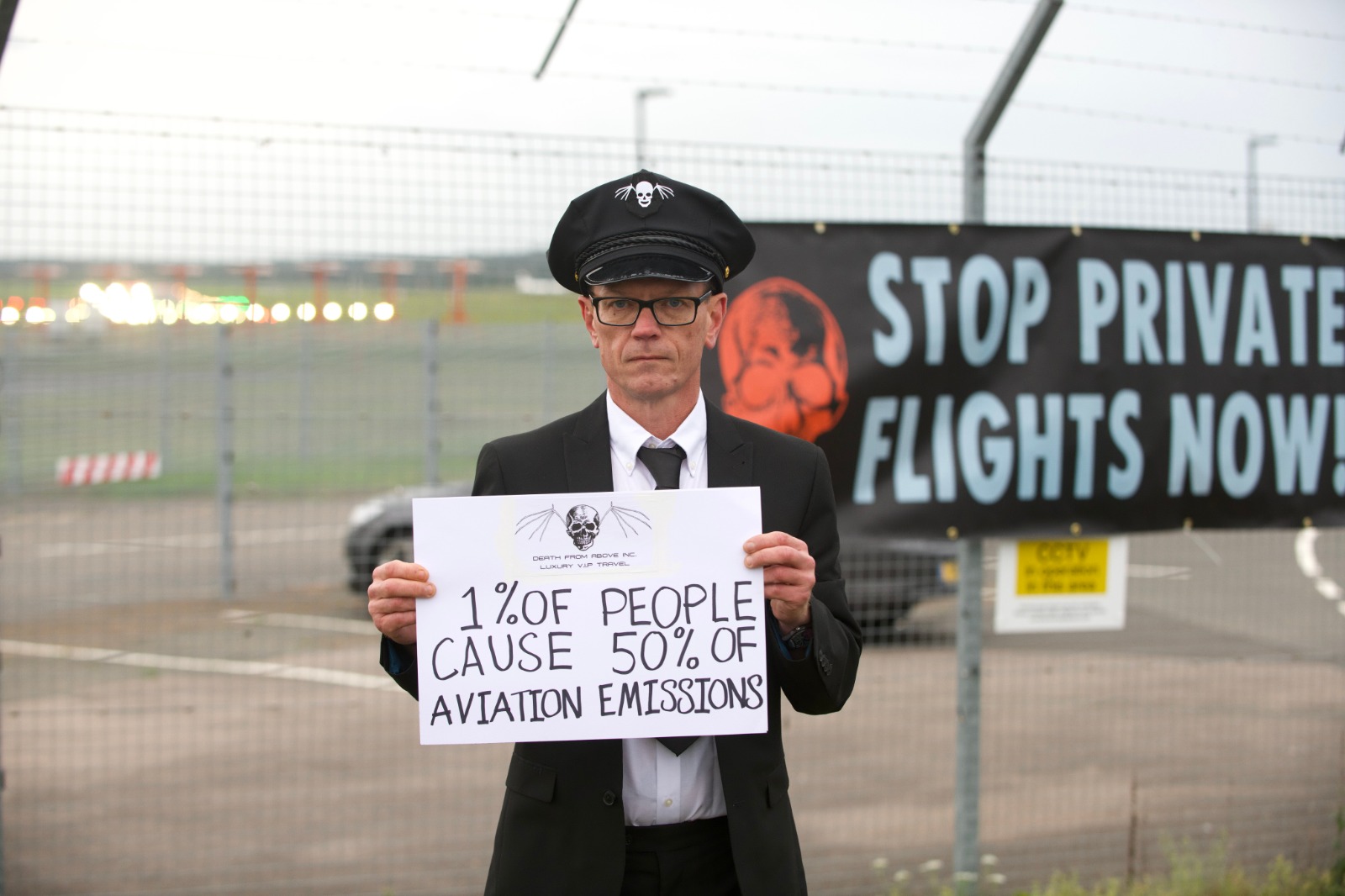 A rebel dressed as a chauffeur holds a sign that says 1% of people cause 50% of aviation emissions.