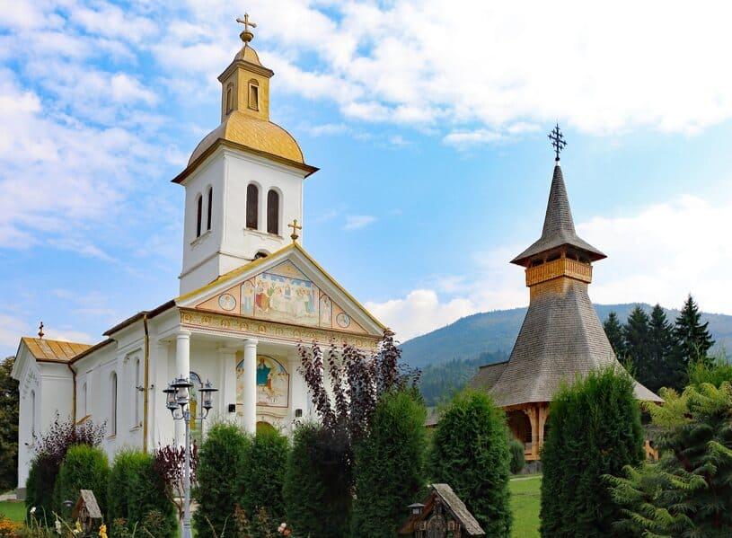 Two wooden churches in Maramures, with mountains in the background and green, lush bushes in the foreground under blue and white skies.