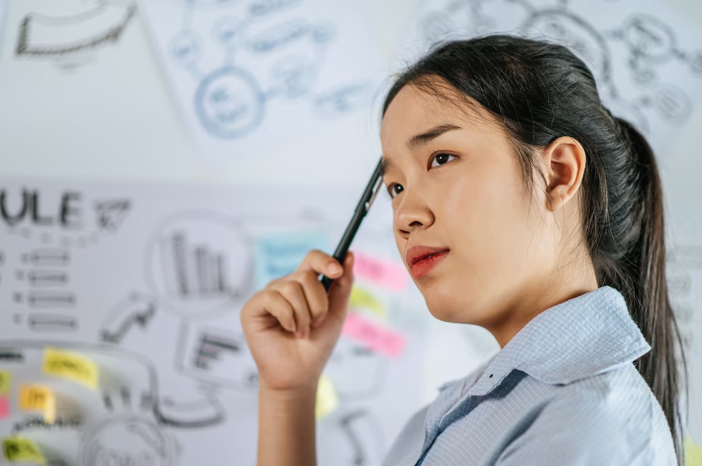 A woman looking into the distance while holding a pen near her head, with a background of a board filled with sketches