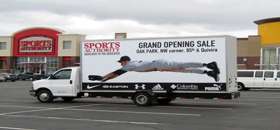 A delivery truck parked at the mall parking lot, prominently featuring billboards for the "SPORTS AUTHORITY" brand. The billboards depict an intense moment of a baseball player diving to make a catch, accompanied by the text "Grand Opening Sale" and the store's address. The truck's sides are adorned with logos of various renowned sports brands, creating a dynamic and enticing advertisement.