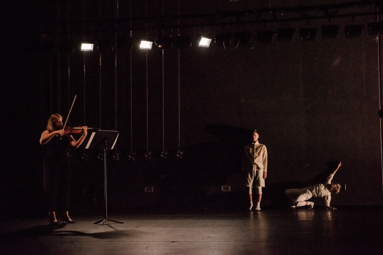 On a darkly lit dance stage, a violist plays on the left while two students dance on the right.