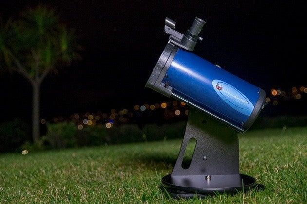 The Astronomers Without Borders OneSky Reflector Telescope, which is blue and black, standing on a grassy field at night with city lights out of focus in the distance.