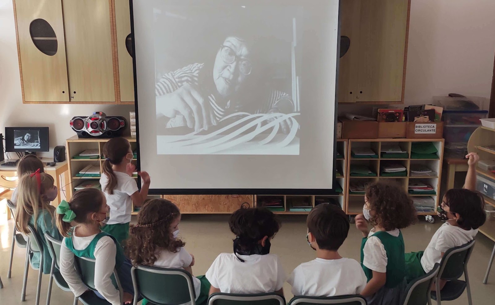 A imagem mostra uma roda de crianças assistindo a uma transmissão em um telão.