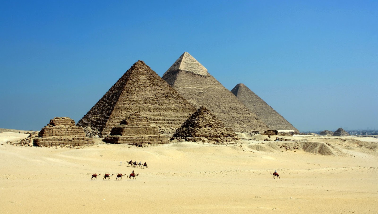  podemos observar na imagem várias das pirâmides do Egito, na frente delas há algumas pessoas andando a camelo