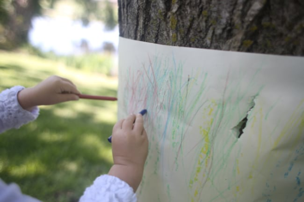 Child coloring with crayons on a piece of white paper wrapped around a thick tree trunk.
