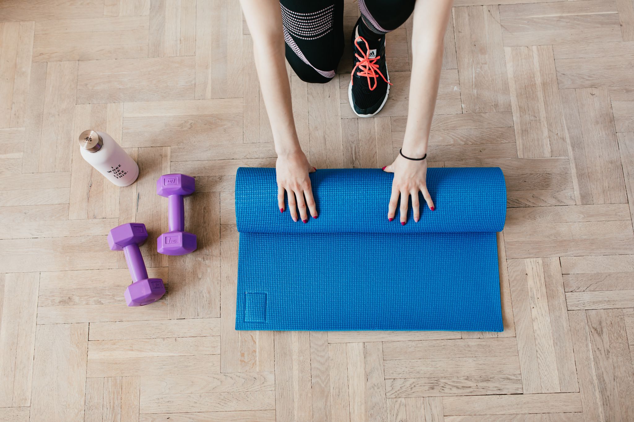 Visão aérea de uma mulher desenrolando um tapete azul de yoga. Ela possui as unhas pintadas de vermelho e veste um tênis preto com cadarços laranjas. Ao lado do tapete, é possível ver duas anilhas roxas e um produto branco, possivelmente um hidratante.