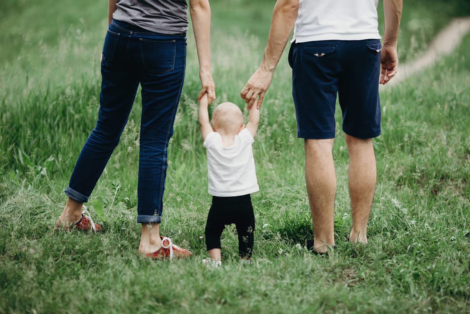 New family, mom, dad, and toddler walking through grassy field. Holding hands with new baby