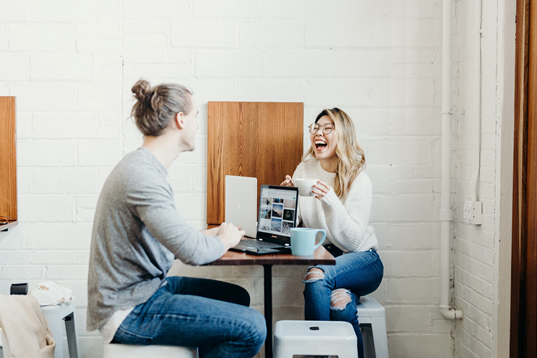 Dos personas en una mesa tomando café y riendo mientras trabajaban desde computadoras portátiles.