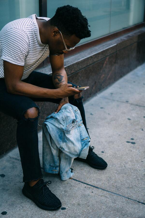 Foto de um homem negro na rua, sentado, segurando uma camisa jeans e mexendo no celular