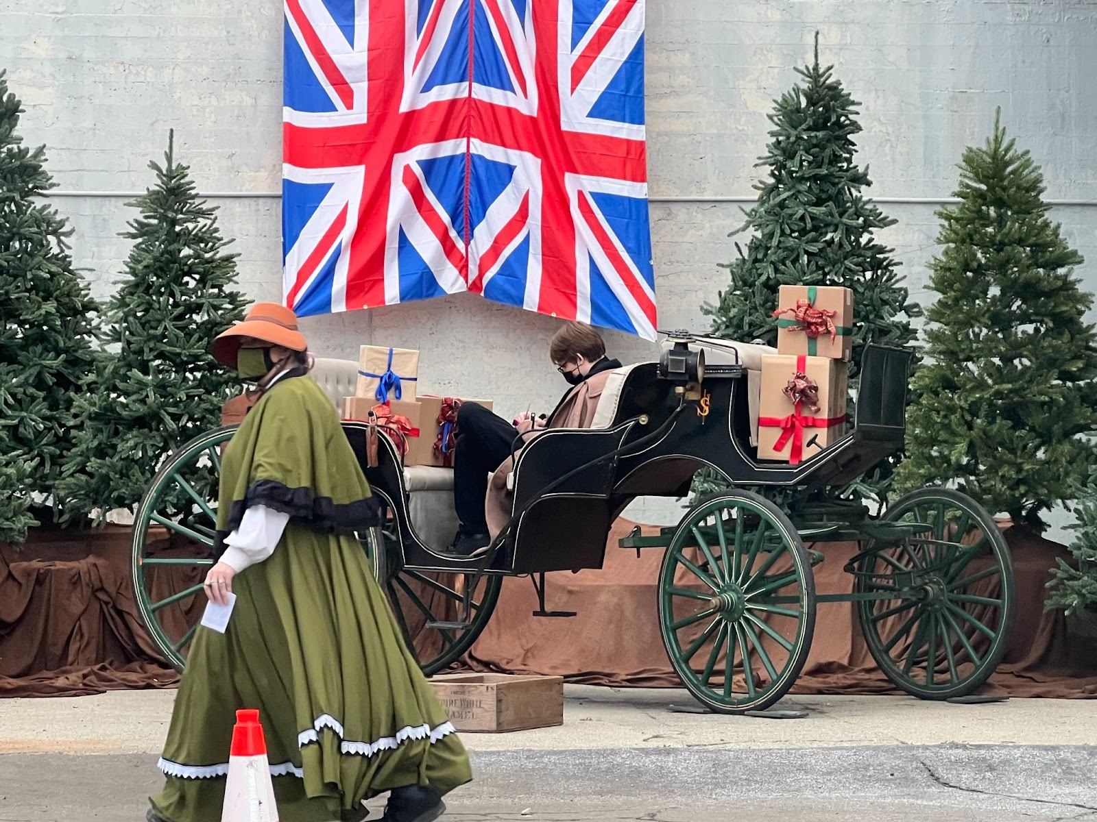 A woman dressed in 19th century British clothing walks in the foreground while a British flag, a carriage, and Christmas trees line the background.