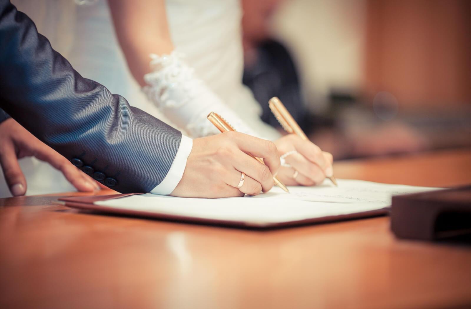 A man's right arm is in a blue colored sleeve and a woman's right hand is in a white wedding glove and both holding a golden coloured pen and signing a marriage contract on the table.