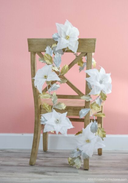 Gold and silver DIY Christmas paper garland tutorial hangs on a light wood chair in front of a salmon-colored wall. 