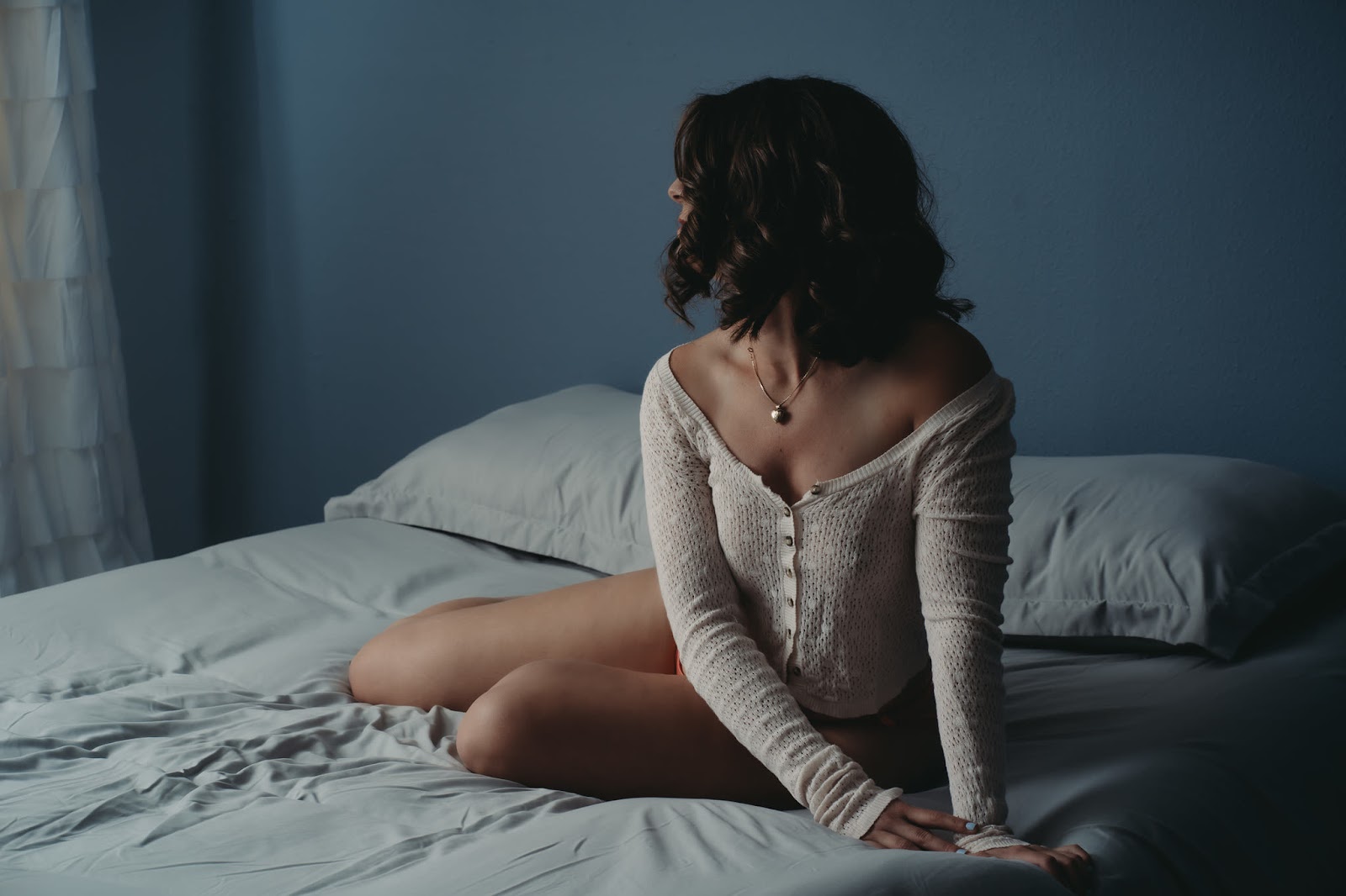 A woman sits on a bed, wearing a gold heart locket and a lacy cardigan that reveals her collarbones and shoulders. Photo by Embodied Art Boudoir. Classic boudoir photography, classic photography, classic boudoir, classy boudoir, classy photography, colorado boudoir, denver boudoir, boulder boudoir, colorado springs boudoir, boudoir ideas, boudoir poses, boudoir inspiration, photography inspiration, boudoir session, boudoir photos