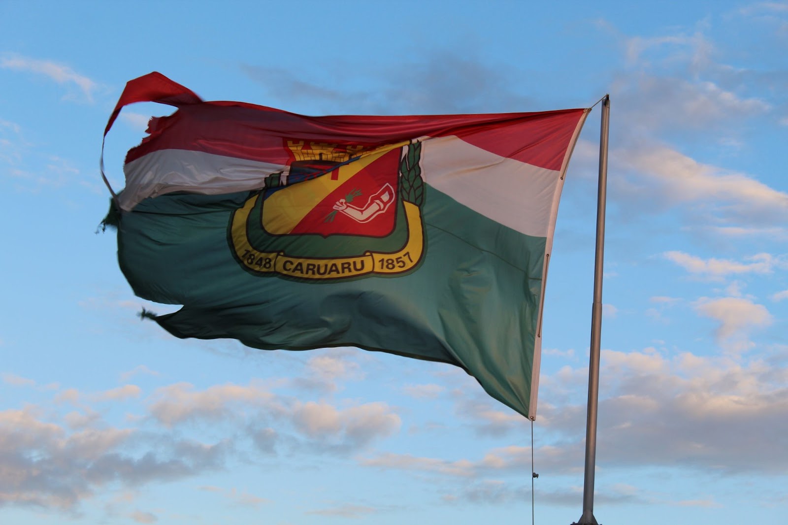 Bandeira de Caruaru pendurada em um mastro e balançando ao vento. Ela exibe três faixas horizontais em vermelho, branco e verde, além do brasão da cidade.