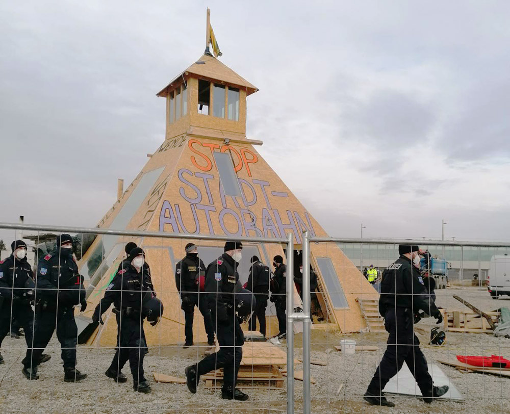 Police storm through a construction site and around a large wooden pyramid structure with Stop The Motorway written on it.