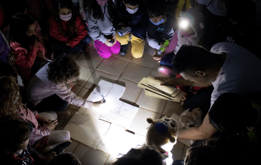 A imagem mostra um grupo de crianças e um adulto observando um mapa com lanternas.