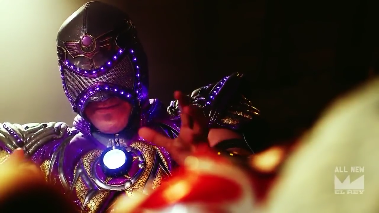 Screencap from Lucha Underground. Aerostar, a luchador wearing a futuristic, astronaut-esque outfit that is predominantly purple and gold with a glowing center, is posed over Fenix, a luchador wearing a red and white bird-like mask. Fenix is resting in a coffin and Aerostar is preparing to revive him.