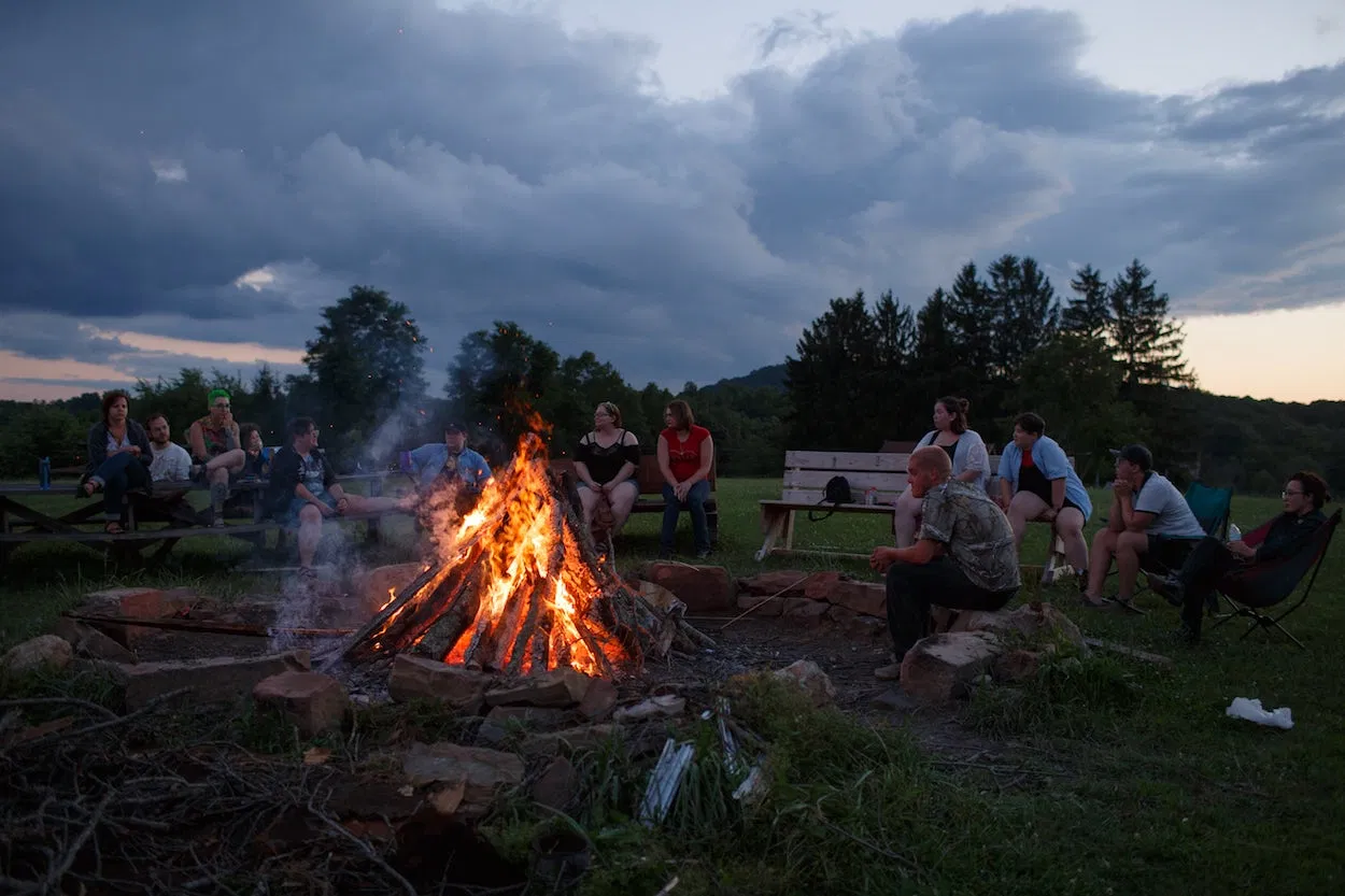 In this picture, a group of people are gathered around a bonfire in an open field lined with trees at dusk.