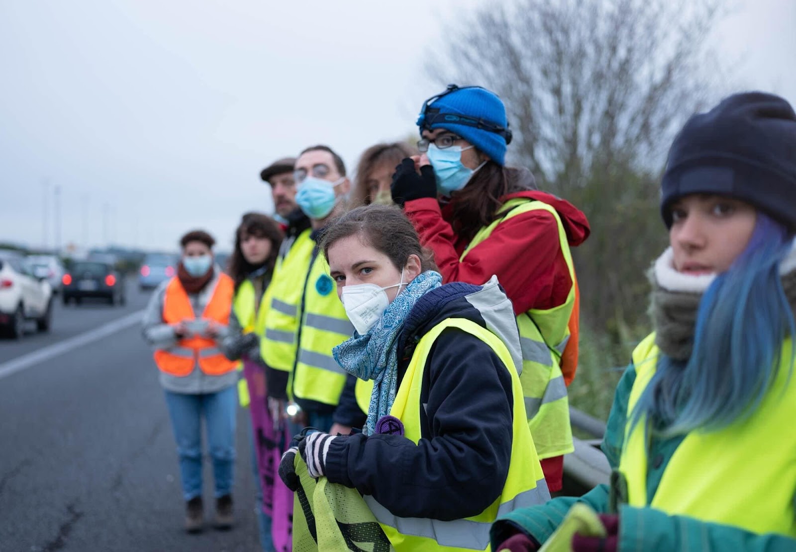 Young rebels in flourescent jackets stand in a motorway layby looking anxiously at the traffic as they wait to swarm it.