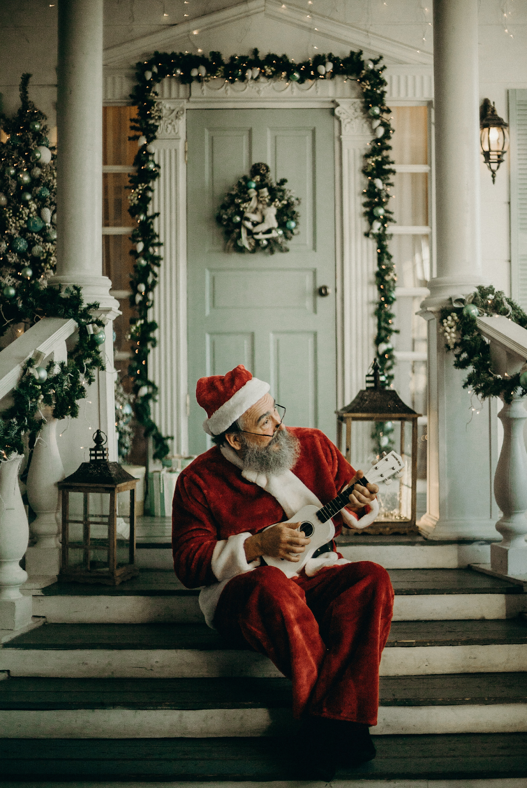 Santa Clause sitting on the porch and playing a small guitar in front of a Christmas decorated house