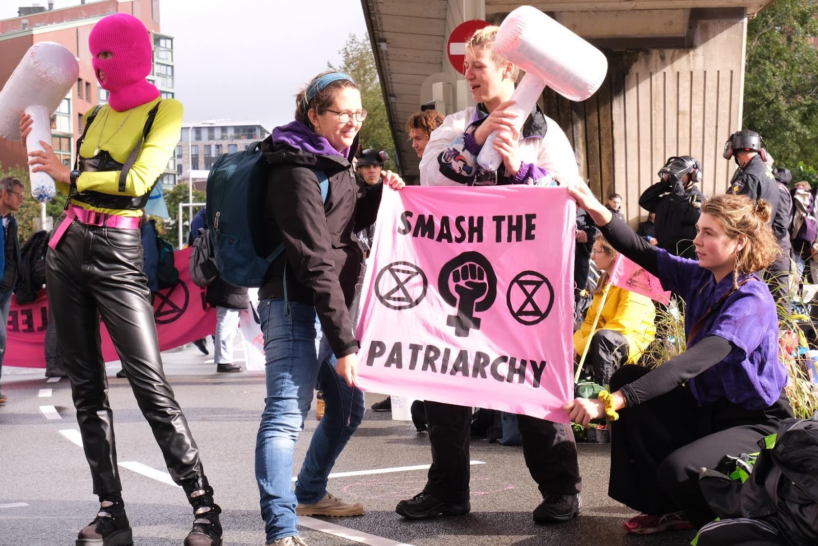 Rebels hold pink hammer shaped cushions and a sign that says smash the patriarchy. Nearby a rebel wears fetish gear and a pink balaclava