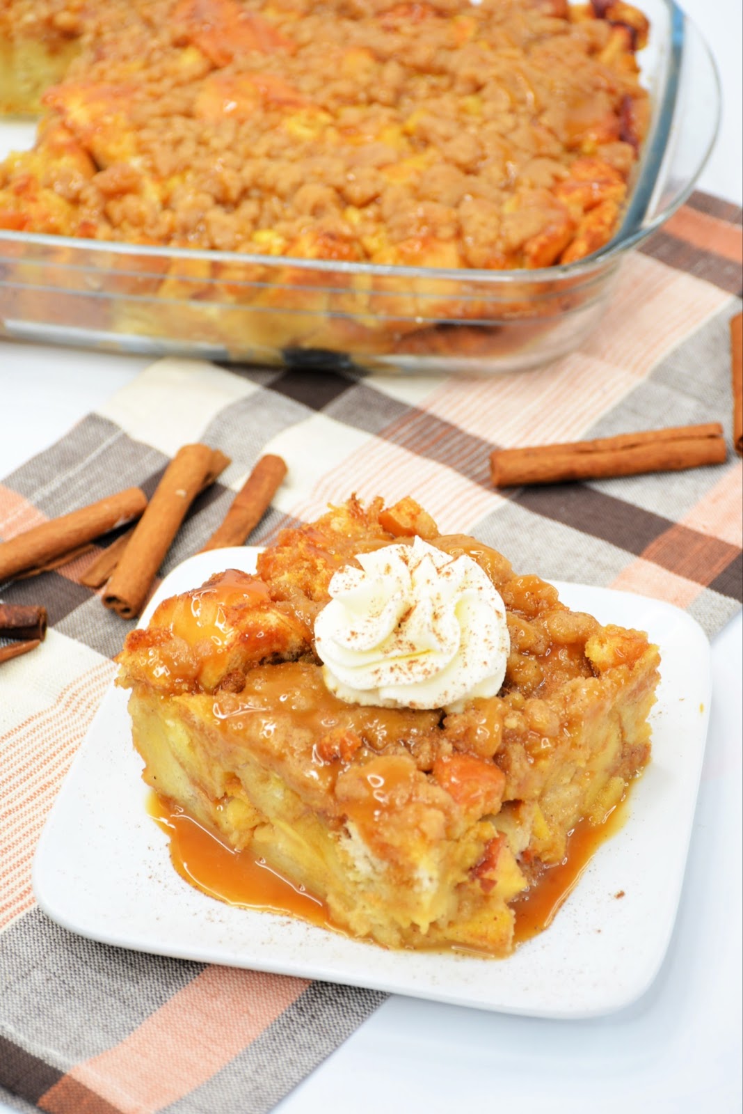 overhead view of slice of baked apple french toast with whipped cream on top on white plate with cinnamon sticks in background 