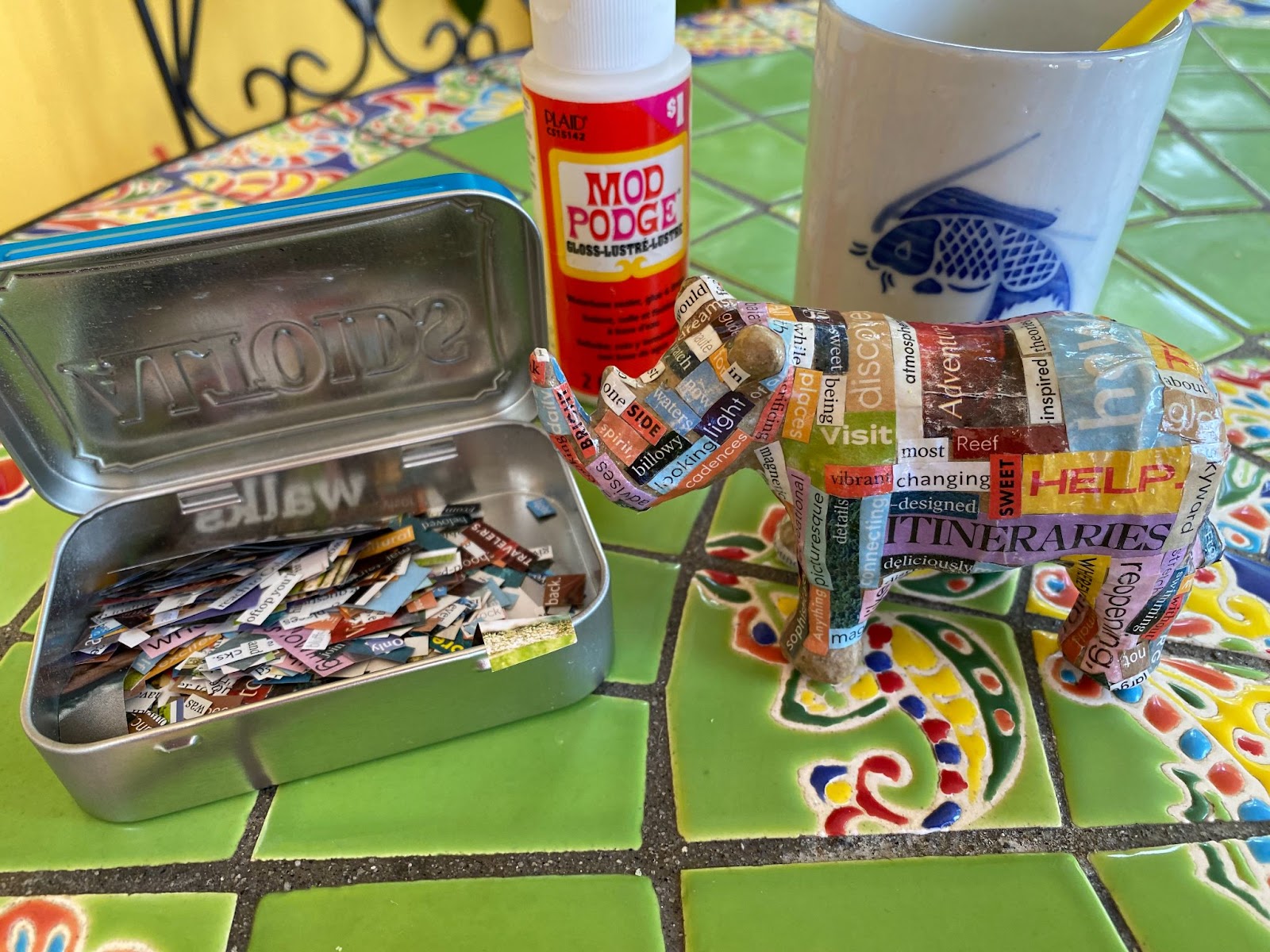 An Altoids tin full of cut out words, a little bottle of Mod Lodge, a cup with a paintbrush, and a pair mace rhinoceros covered in word collage.