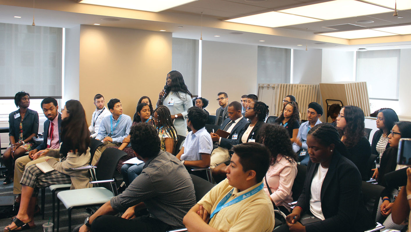 A group of medical interns in scrubs sitting in a classroom, asking questions during a training session.