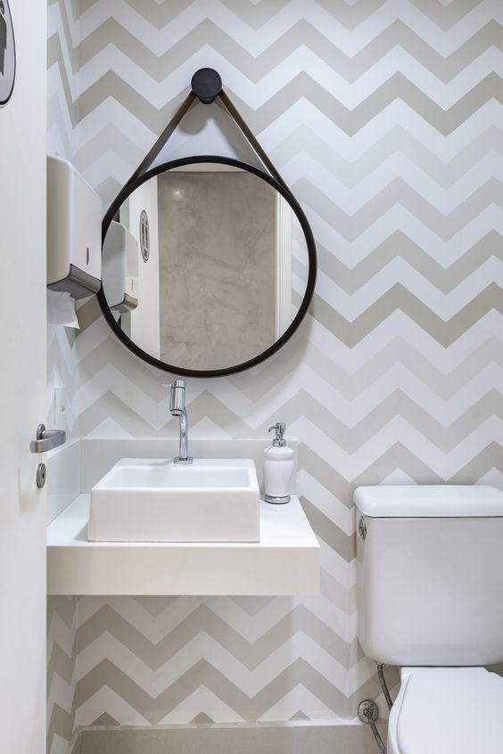 Lavabo em estilo clean, com papel de parede desenhado em tons claros, louças brancas e espelho redondo.