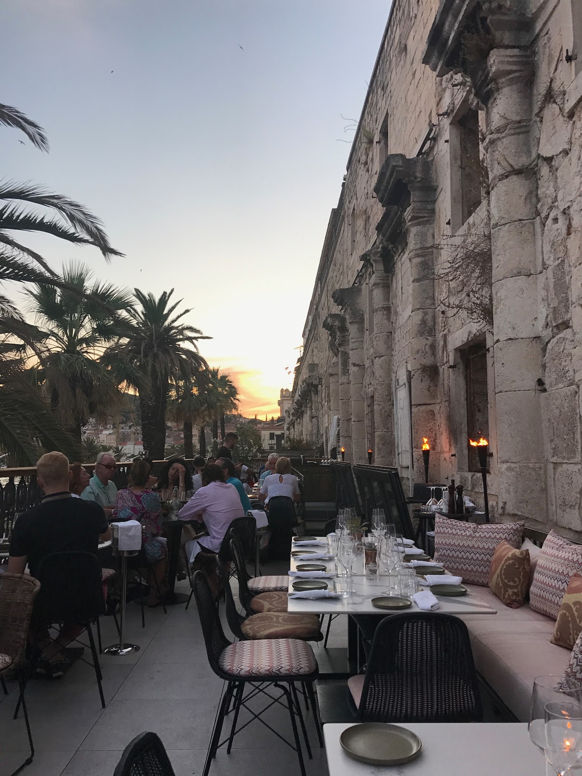 Prestigious outdoor restaurant by the walls of Diocletian's Palace. Tourists, outdoor terrace next to palm trees and one large empty table. beautiful sunset in split croatia