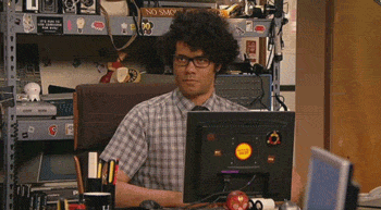 A scene from the TV show 'IT Crowd', Moss is sitting as his desk and grabs a bucket of popcorn and proceeds to eat it whilst looking forward. 